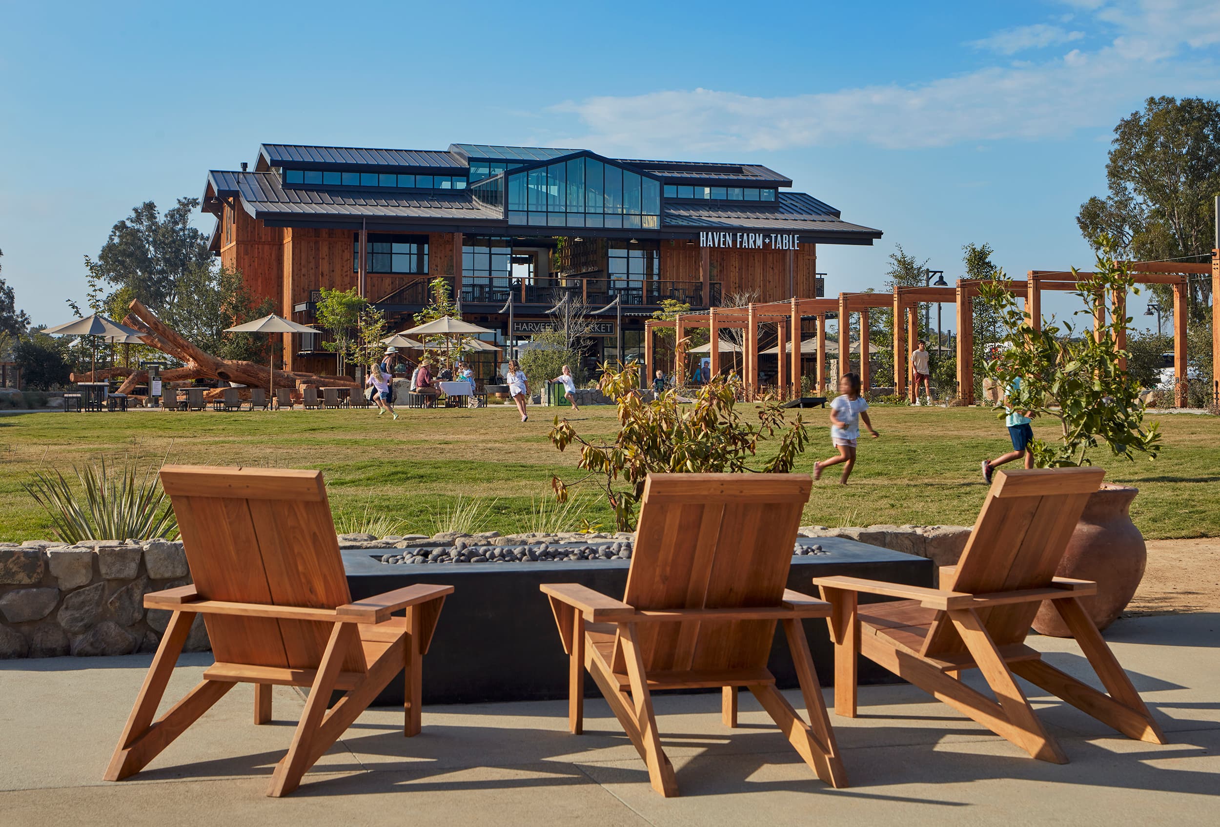 Haven Farm + Table at Fox Point Farms on a clear afternoon: three timber lounge chairs around a fire pit on the lawn, children running across the grass, and the glass-gabled barn behind