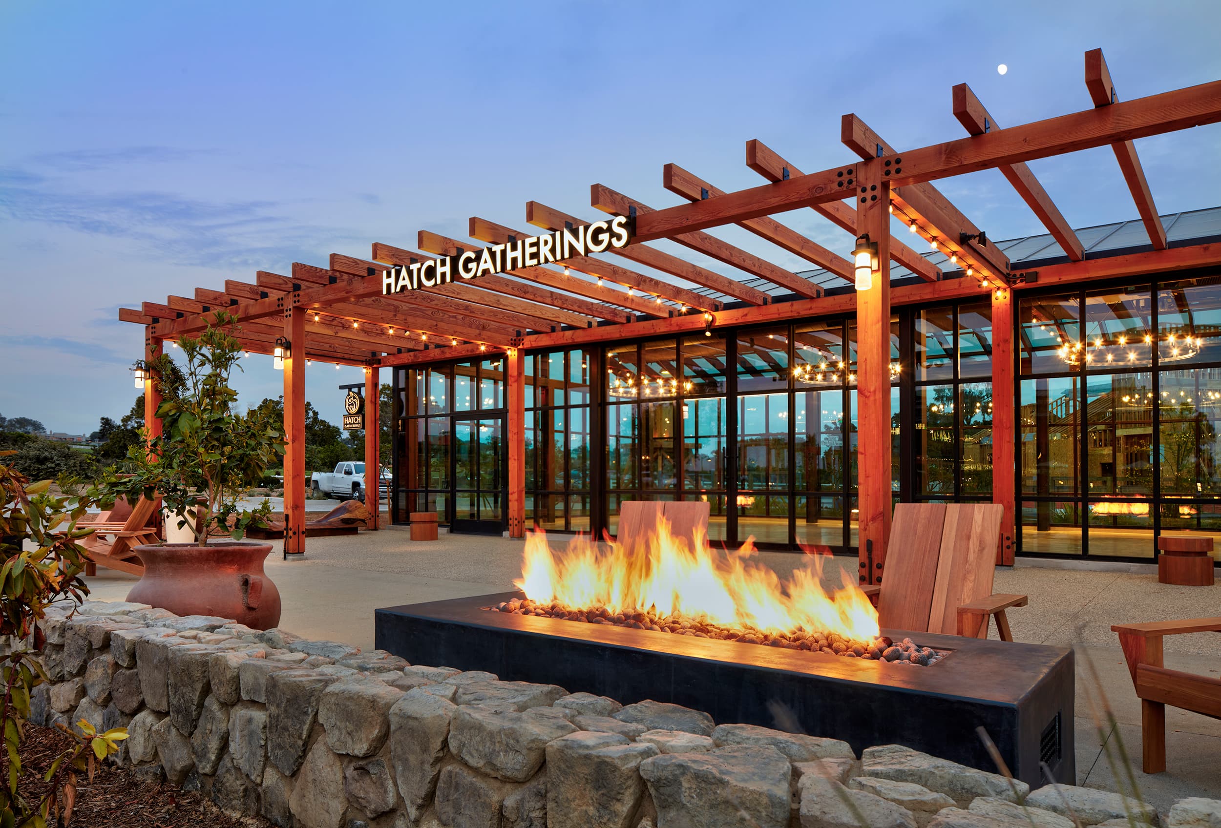 Hatch Gatherings at Fox Point Farms in North County San Diego at dusk: a timber pergola strung with lights over a glass-walled events hall, a long fire pit burning before it behind a stone wall, and the moon above