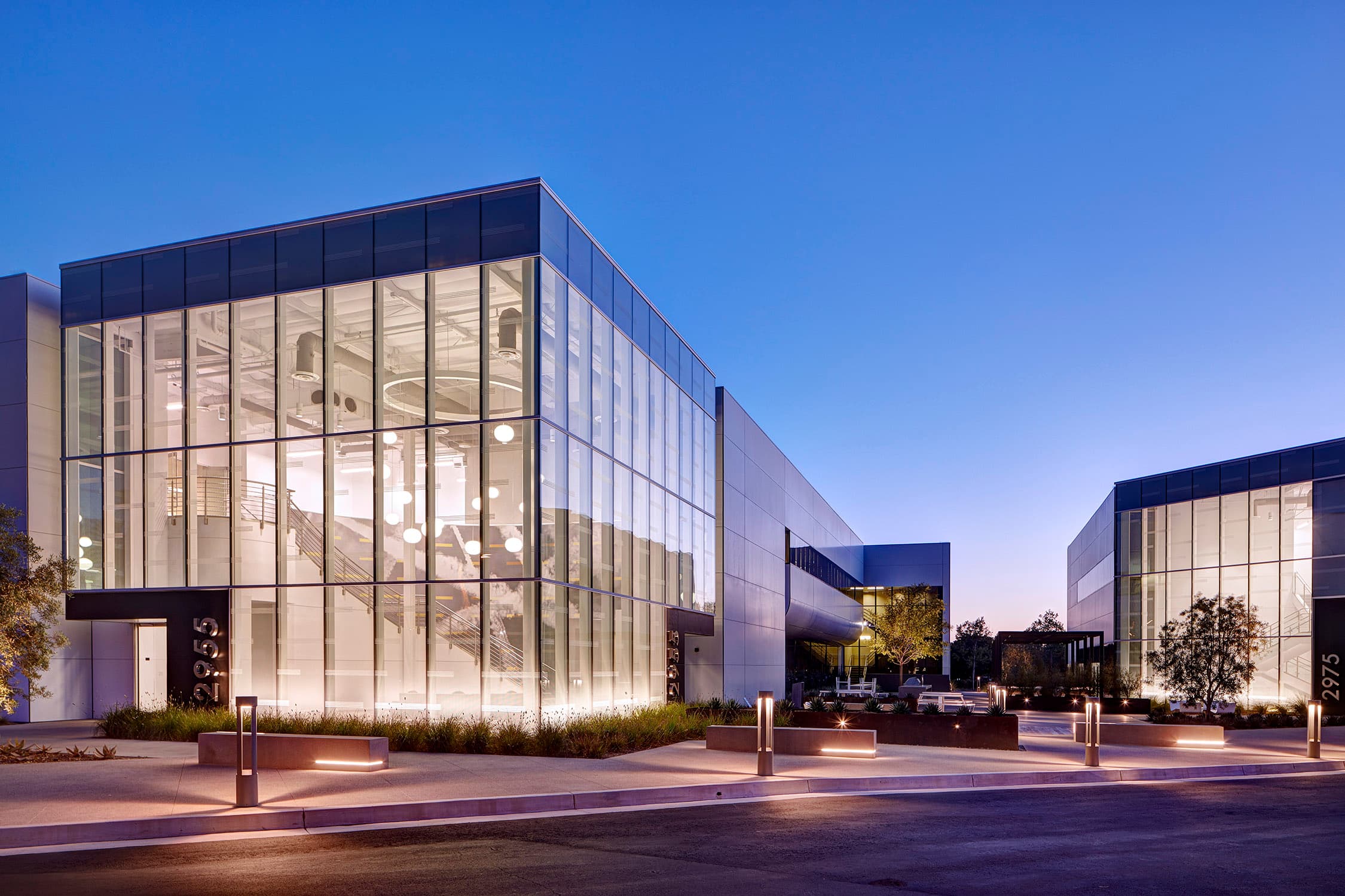 Landing at Red Hill at dusk: a two-storey fritted glass entry pavilion glowing from within at the corner of an office campus, lit concrete benches and bollards along the drive, and a matching pavilion across the courtyard