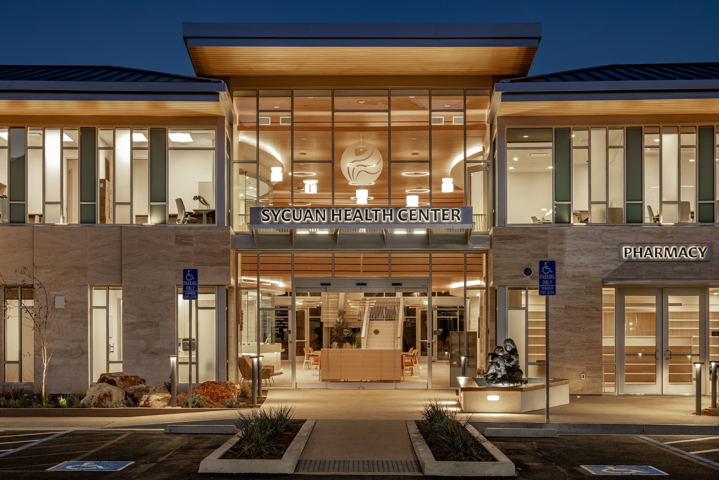 Sycuan Health Center in El Cajon at dusk, a two-storey stone and glass entry lit from within under a timber canopy