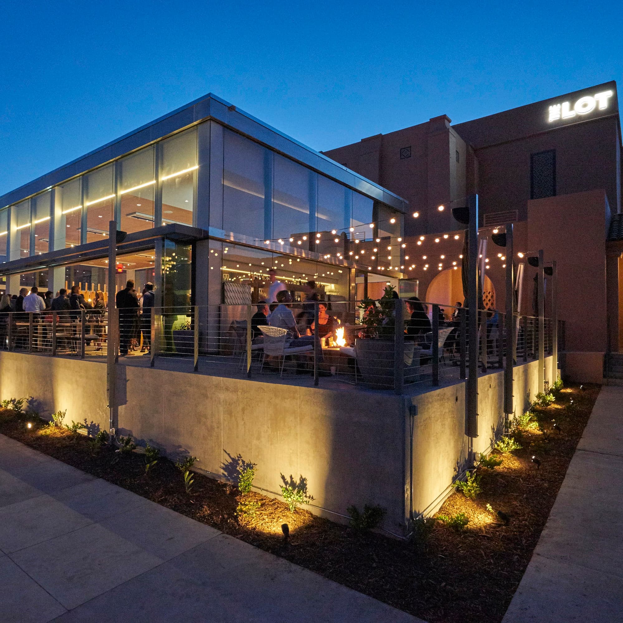 The Lot at Liberty Station at dusk: a crowd on the terrace of a glass-walled bar under strings of lights, gathered around a fire table, with the Lot sign on the building behind